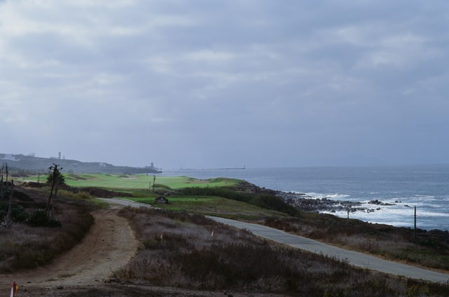 A cart path runs through coastal scrub beside the fairways and rocky shoreline.