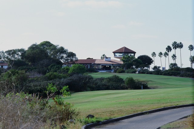 A building with red tile roofs and a glass tower stands beyond the fairway, trees, palms, and cart path.