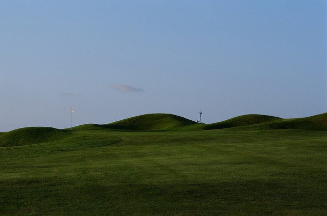 Rounded grassy mounds surround a white flag beneath a blue sky.