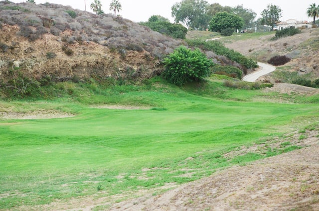 The maintained former eighteenth green sits in a rocky hollow beneath a winding cart path.
