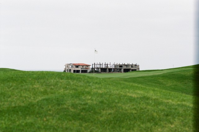 A white flag stands on a green with unfinished buildings and exposed columns behind it.