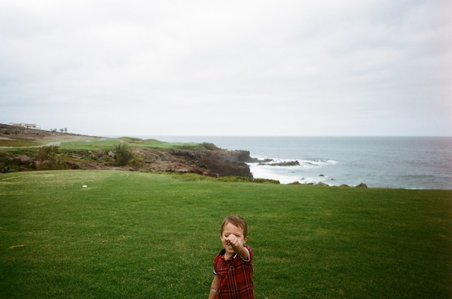 Baker points toward the camera from the grass, with rocky cliffs and the ocean behind him.