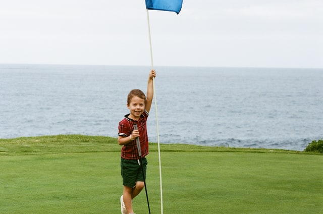 Baker holds a blue flagstick and a putter on a green above the ocean.