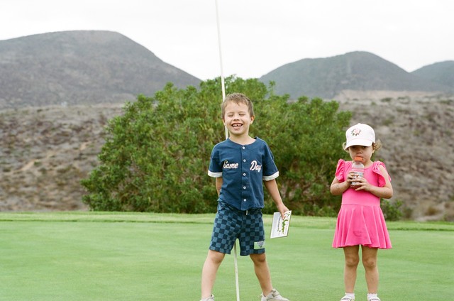 Baker smiles beside Romee, who holds a plastic bottle, on a green backed by dry hills.