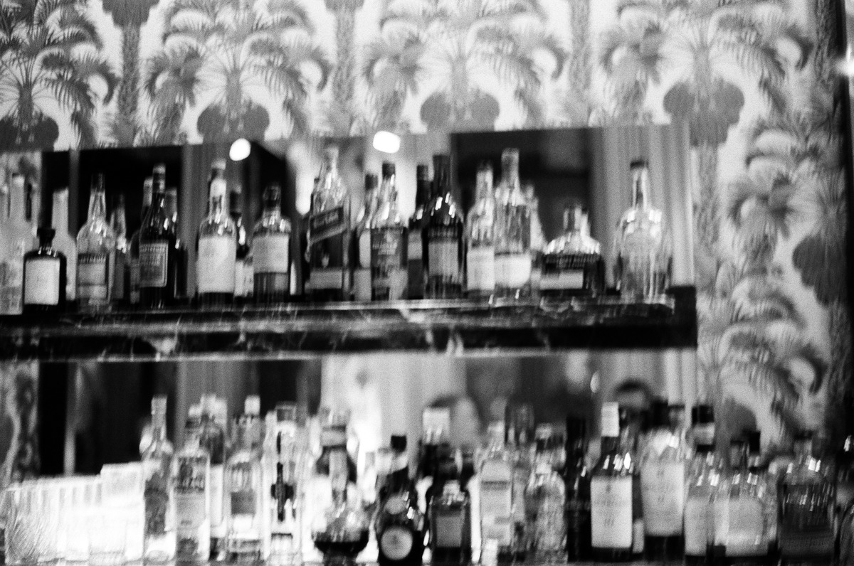 A softly blurred black-and-white photograph of bar bottles and palm-patterned wallpaper