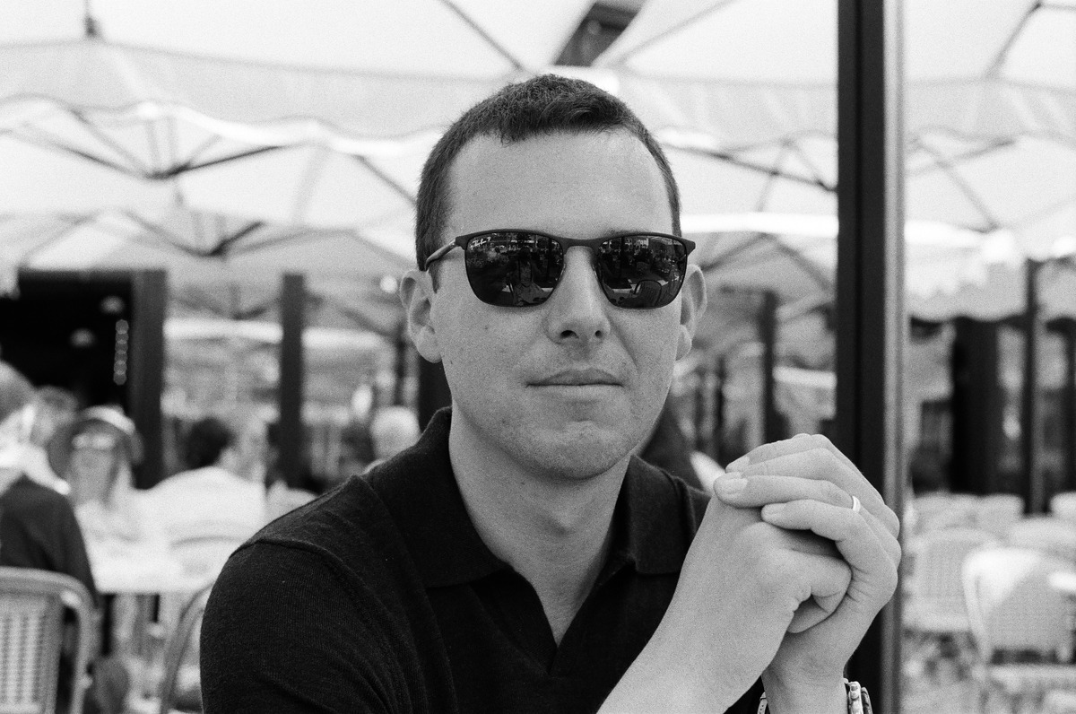 A man in sunglasses seated at a table, with his hands clasped, in black and white