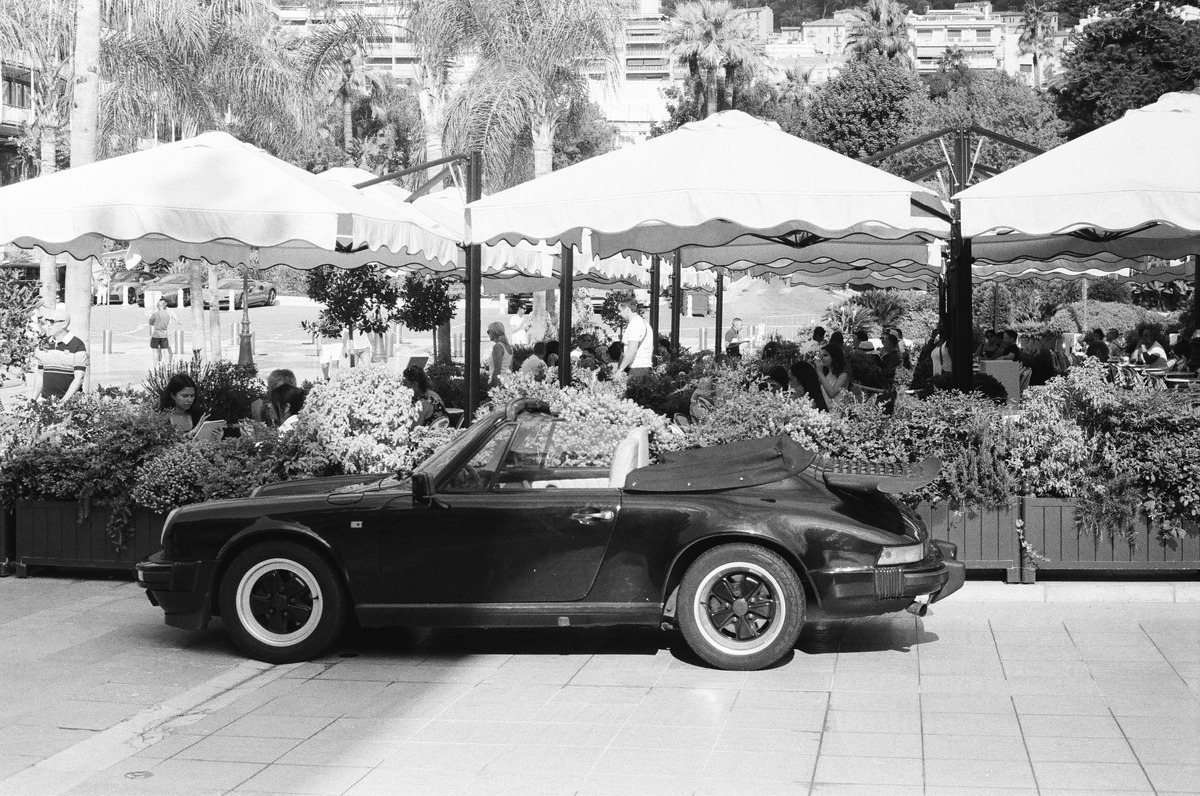 A black-and-white photograph of a convertible parked outside a busy café