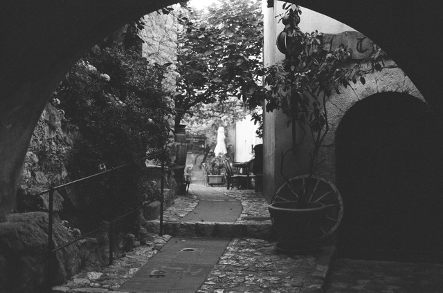 A black-and-white photograph of a stone arch and path beside a pot and wagon wheel