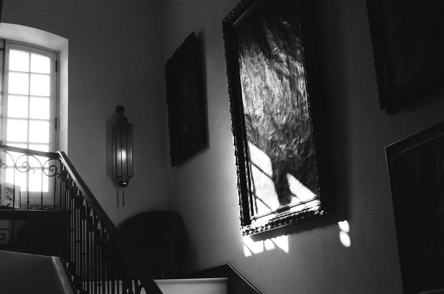 Window light falling across framed artwork beside a staircase, in black and white