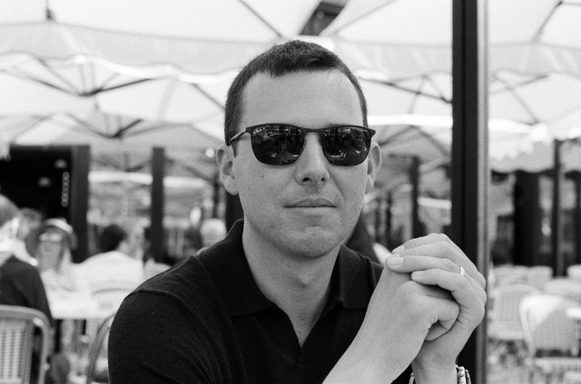 A man in sunglasses seated at a table, with his hands clasped, in black and white