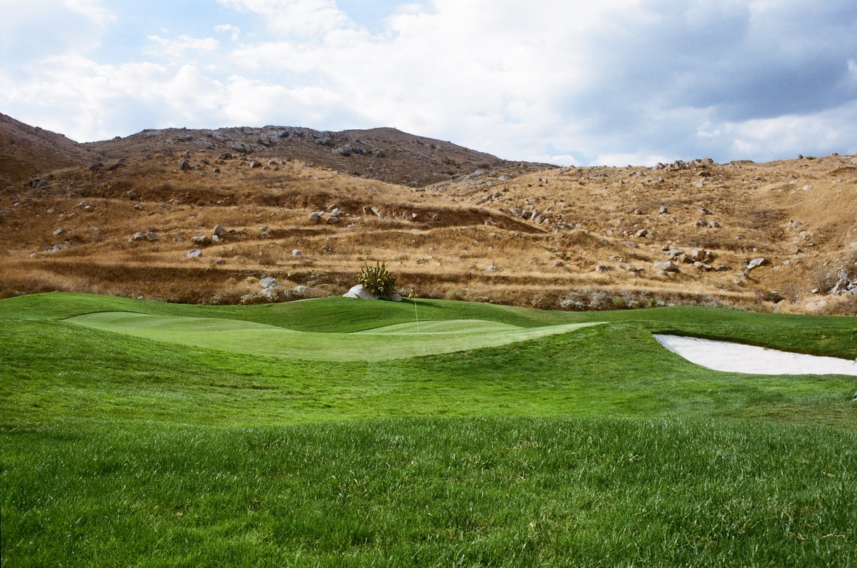 A contoured green and white sand bunker beneath a dry hillside scattered with boulders