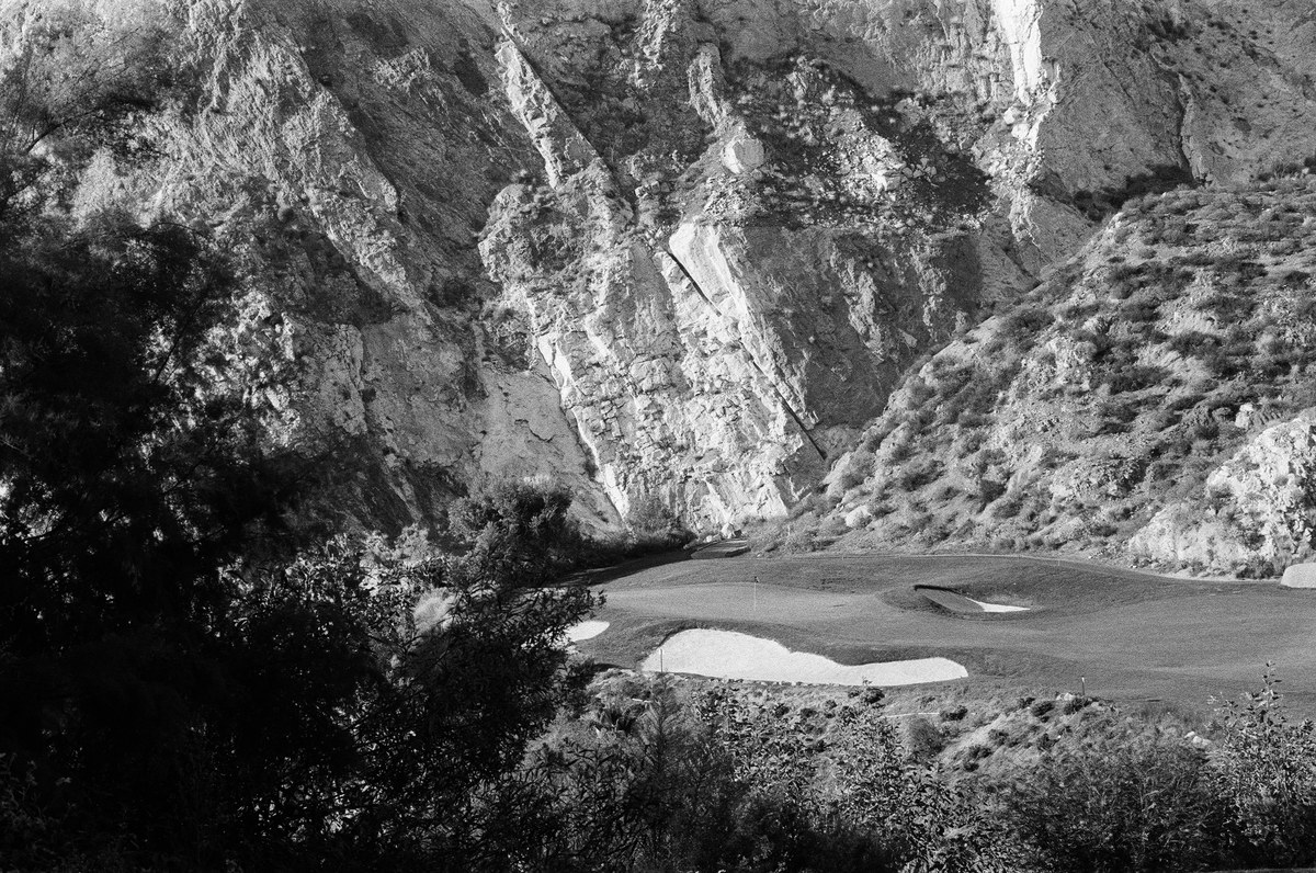 Black-and-white view of the green and bunkers beneath a textured quarry wall, framed by a dark tree at left