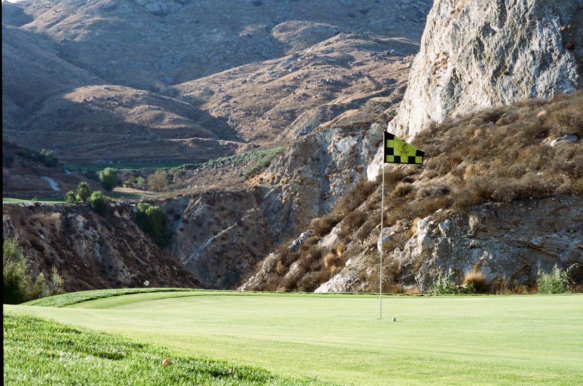 A golf ball near a yellow and black flag on a green, with quarry walls and hills behind