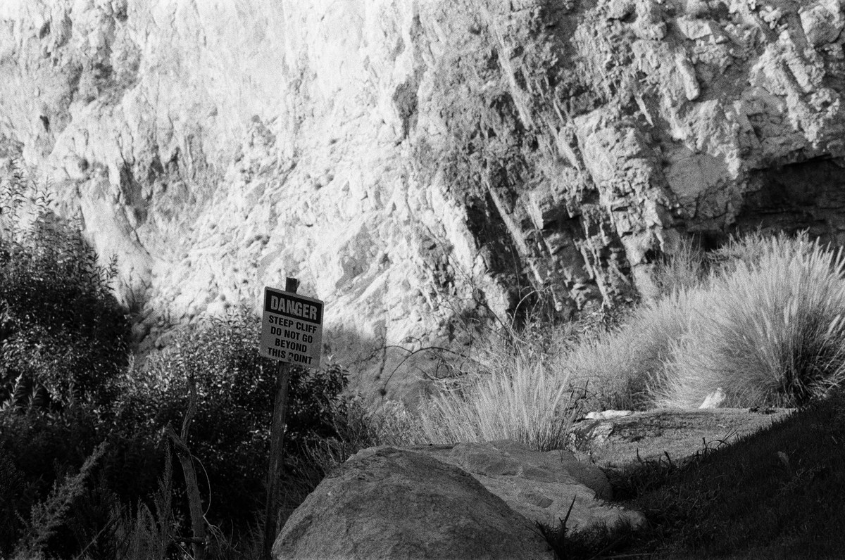 Black-and-white photograph of a steep-cliff warning sign among grasses and rocks against the quarry wall