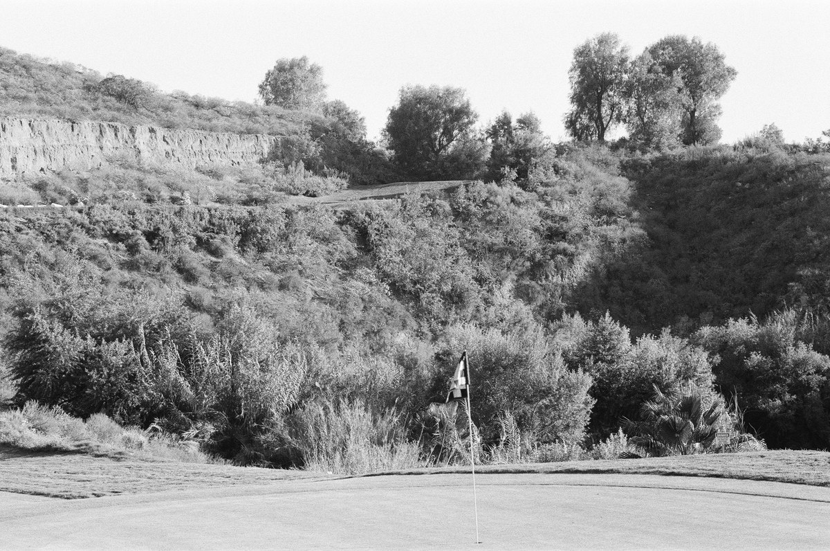 Black-and-white photograph of a flag on a green, with dense vegetation, rock ledges, and trees behind